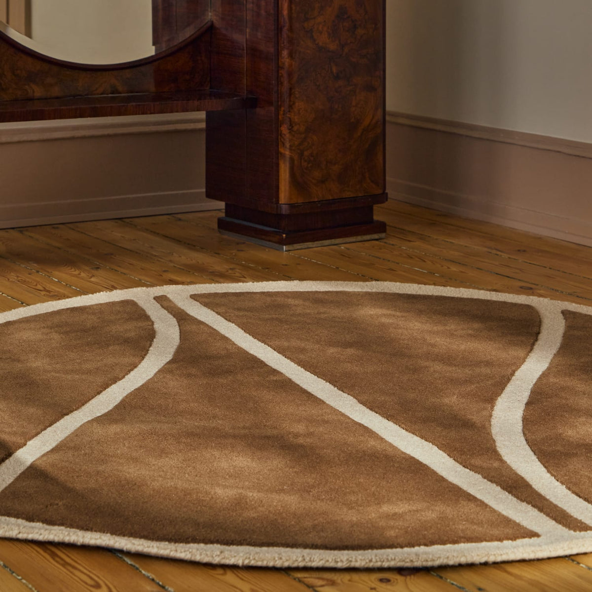 A round area rug with a brown and beige basketball-style court design, shown on a wooden floor in a room with a dark wood furniture piece in the background.