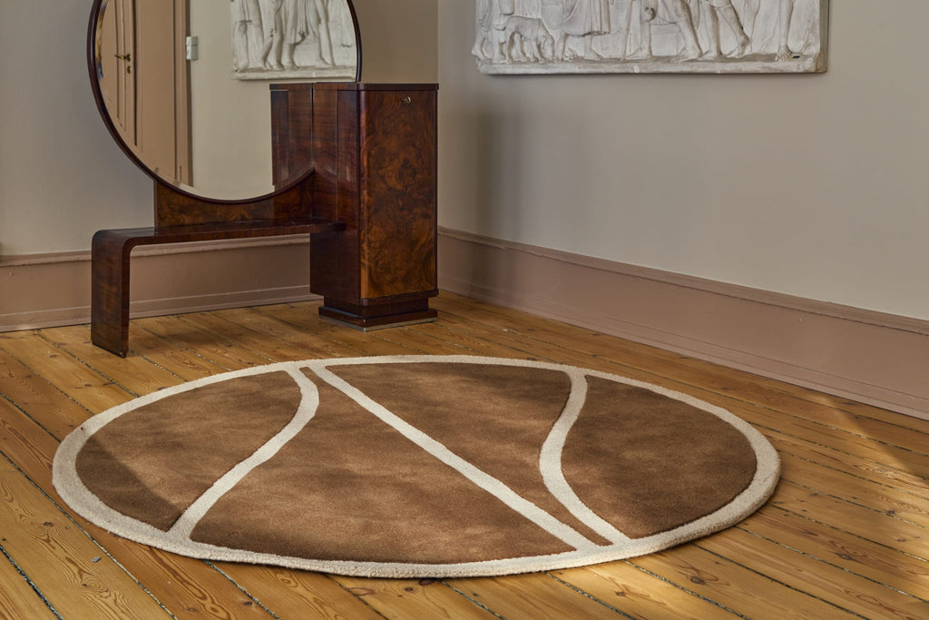A round area rug shaped like a basketball, shown on a wooden floor in a styled room with a wooden bench, mirror, and wall art.