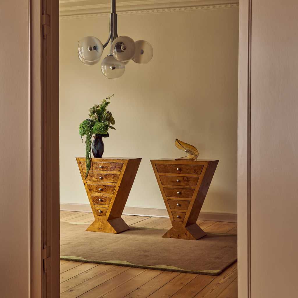 Two matching wooden side tables with geometric V-shaped bases and multiple drawers, styled in a warm, elegant room with a chandelier, rug, and vase of flowers.