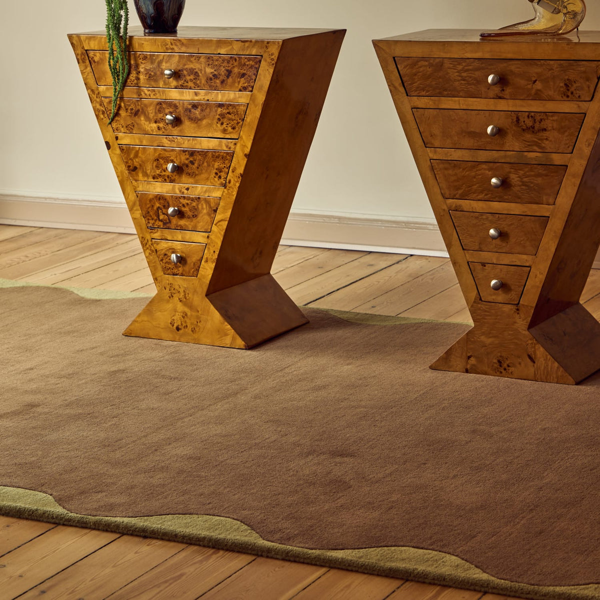 Two matching wooden side tables with angled triangular bases and five drawers each, shown in a styled room setting on a brown area rug with a wood floor.