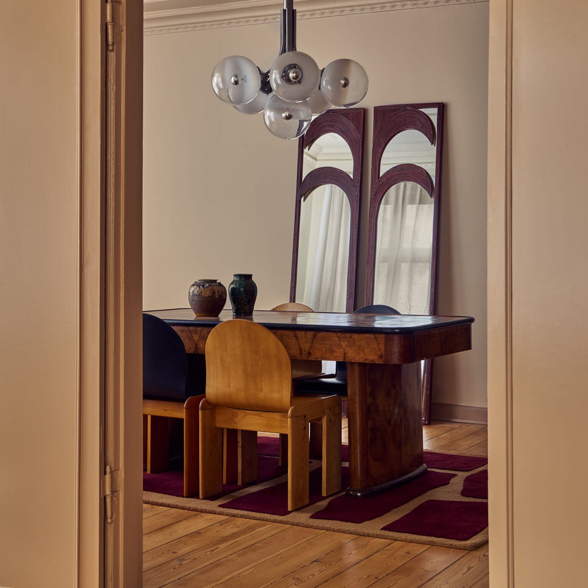 A dining room interior viewed through an open doorway, featuring a dark wood dining table, wooden chairs, a hanging globe chandelier, and a tall decorative mirror with arched glass panels.