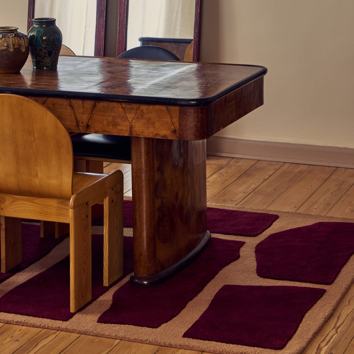 A styled interior scene showing a wooden dining table and chair on a patterned area rug over a wood floor, with a mirror and vases in the background.