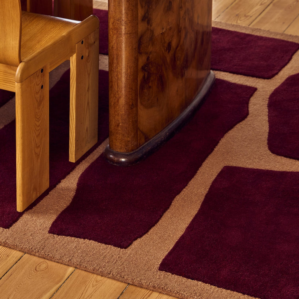 A close-up of a plush area rug with a geometric pattern in deep burgundy and tan, styled in a home setting next to a wooden chair and table leg on a hardwood floor.