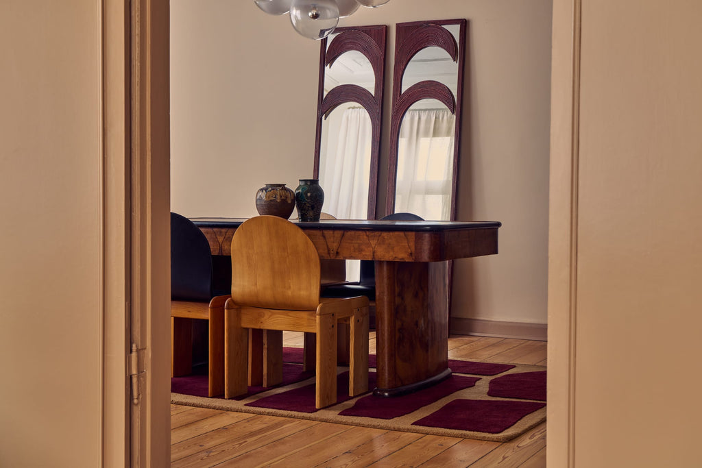 A mid-century modern dining room scene viewed through an open doorway, featuring a dark wood table, a wooden chair, a patterned burgundy rug, and two tall arched mirrors against beige walls.