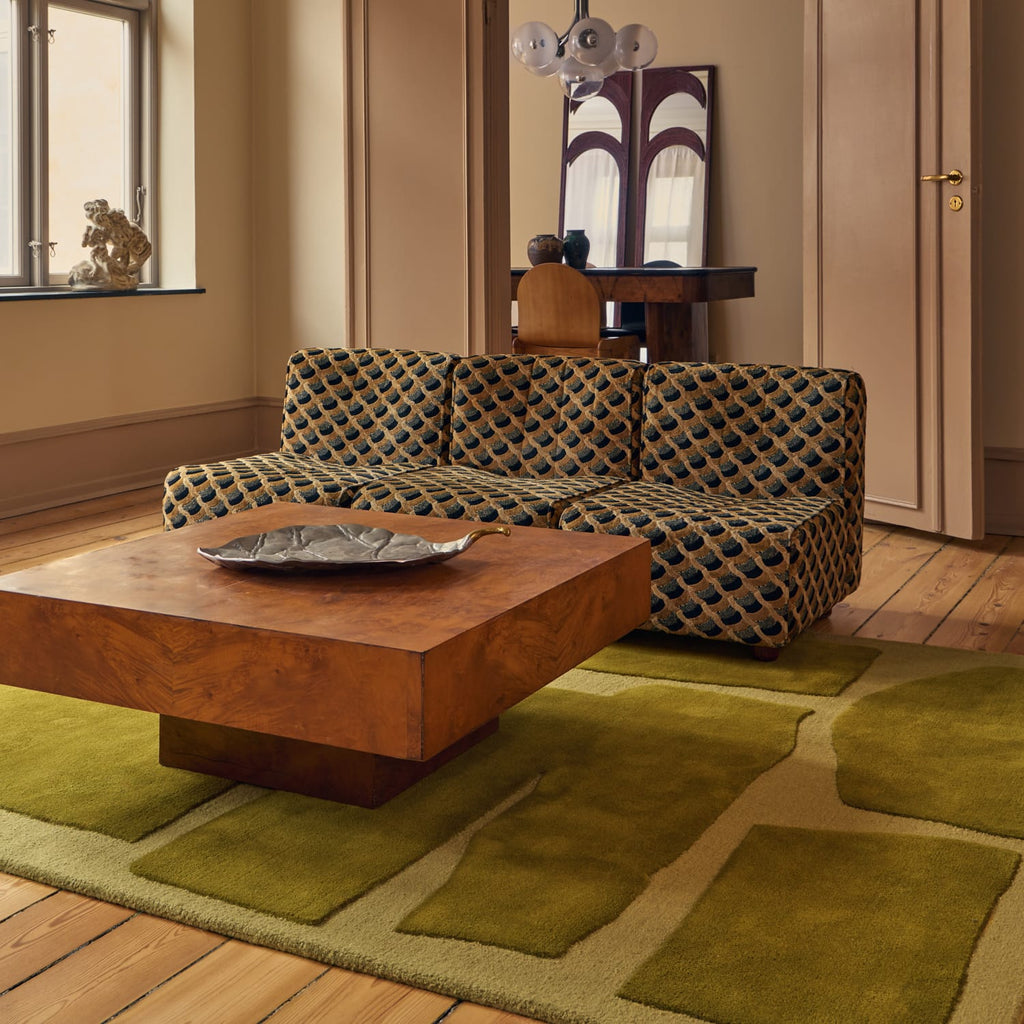 A patterned upholstered sofa in a styled living room with a wooden coffee table, olive green area rug, wood floors, and a window in the background.