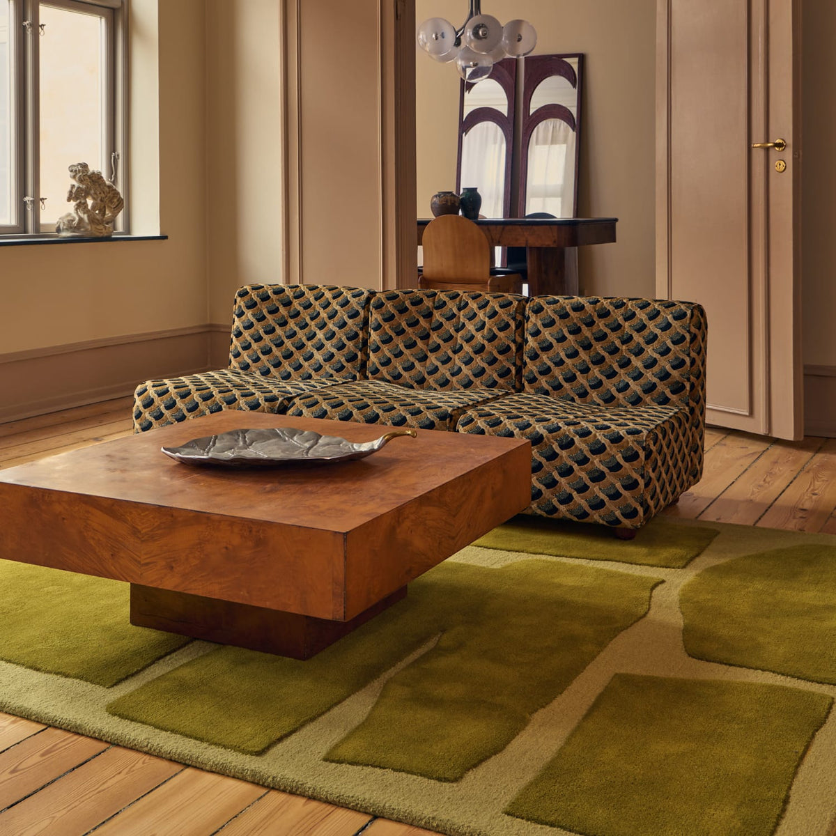 A patterned upholstered sofa in a styled living room with a wooden coffee table, olive green area rug, wood floors, and a window in the background.