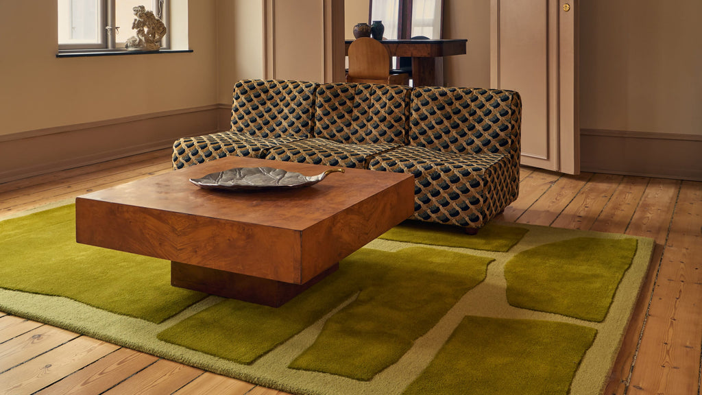 A modern living room scene featuring a large olive green geometric area rug under a square wooden coffee table, with a patterned sofa in the background.