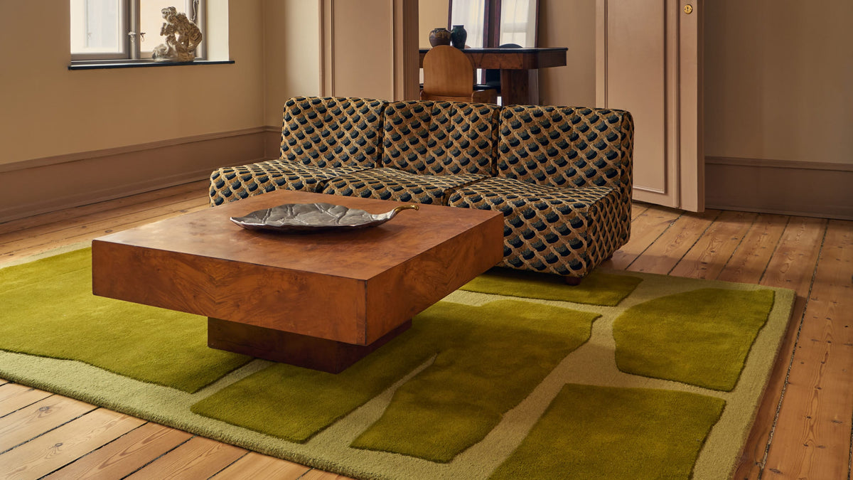 A modern living room scene featuring a large olive green geometric area rug under a square wooden coffee table, with a patterned sofa in the background.