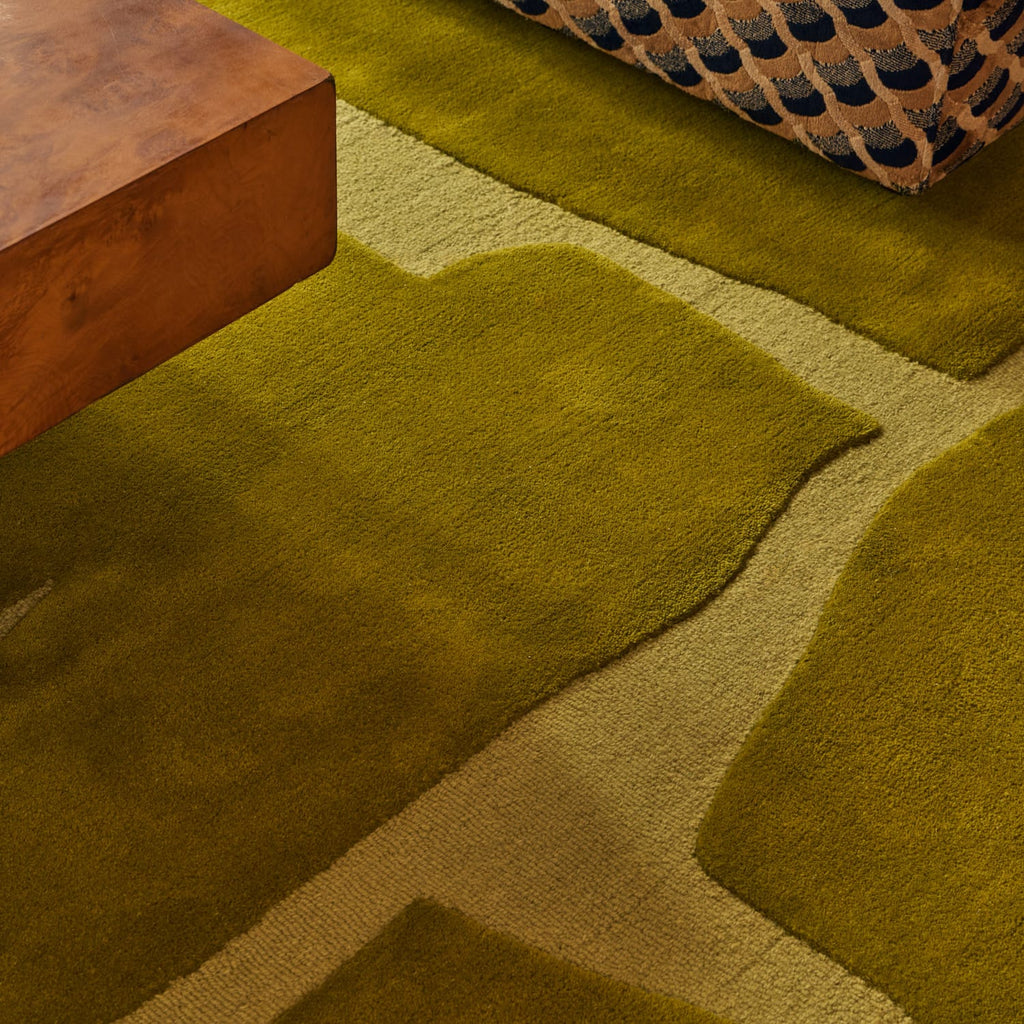 A close-up of a plush area rug with an abstract organic pattern in olive green and beige, styled in a modern interior with a wooden side table and a patterned cushion.