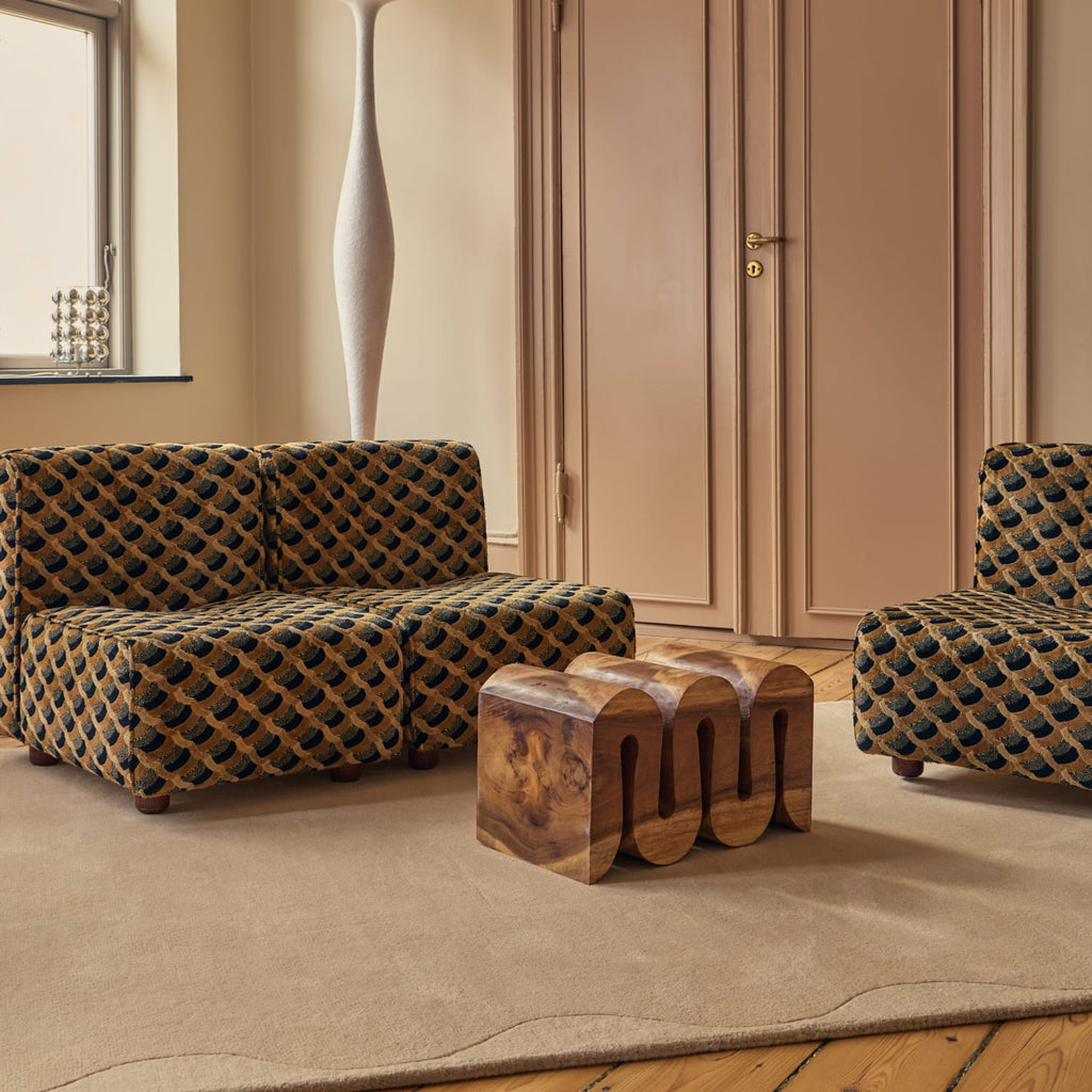 A styled interior scene showing patterned upholstered furniture in a warm room: a low sofa, part of a matching armchair, and a sculptural wooden side table on a beige rug. No people or animals are present, but this is still not a clean isolated product shot because a real room environment is visible.