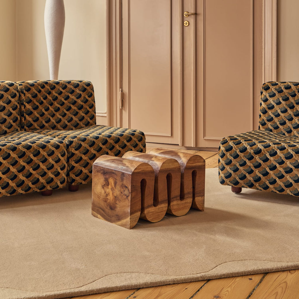 A modern wooden three-seater stool with a curved, wavy backrest sits on a beige area rug in a living room setting, flanked by two patterned armchairs.