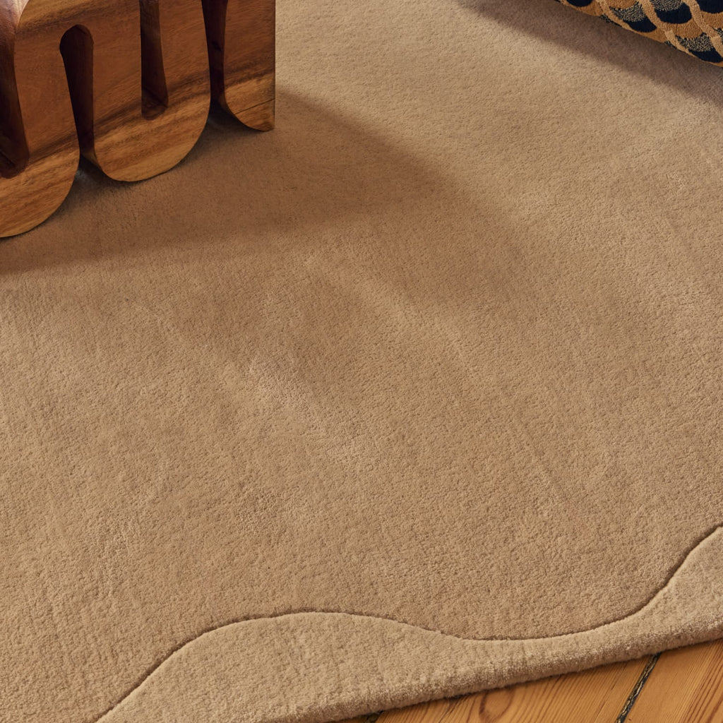 A close-up of a beige textured area rug with a wavy stitched edge, shown on a wooden floor with a wooden furniture leg visible in the top left corner.
