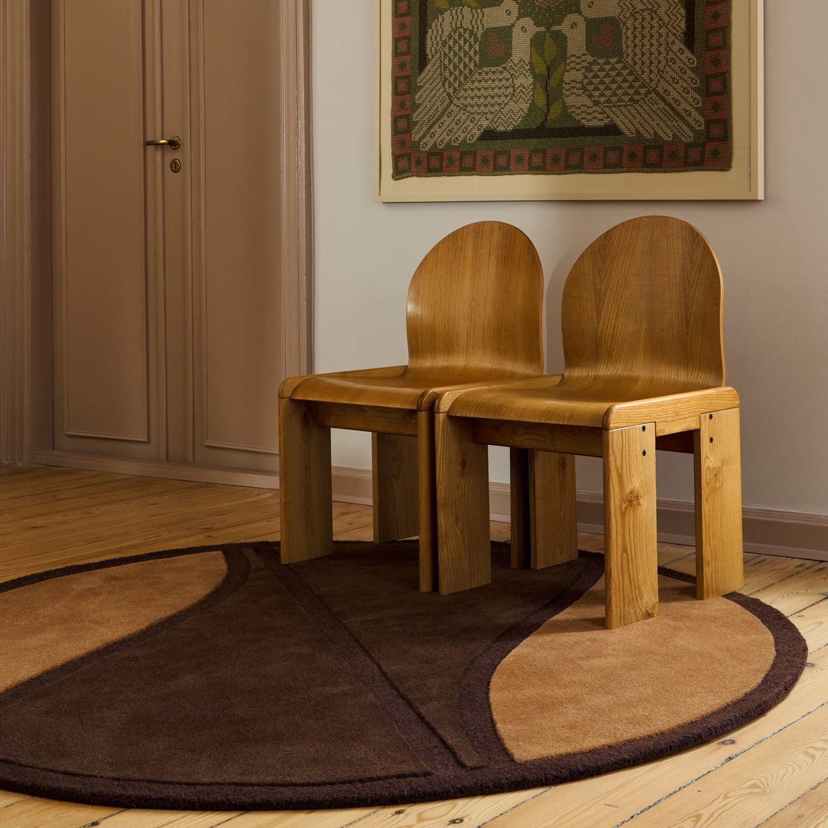 Two light brown wooden chairs with arched backs sit on a round area rug with a dark brown and beige abstract design. The chairs are placed on a light wood floor against a beige wall, with a framed artwork hanging above them.