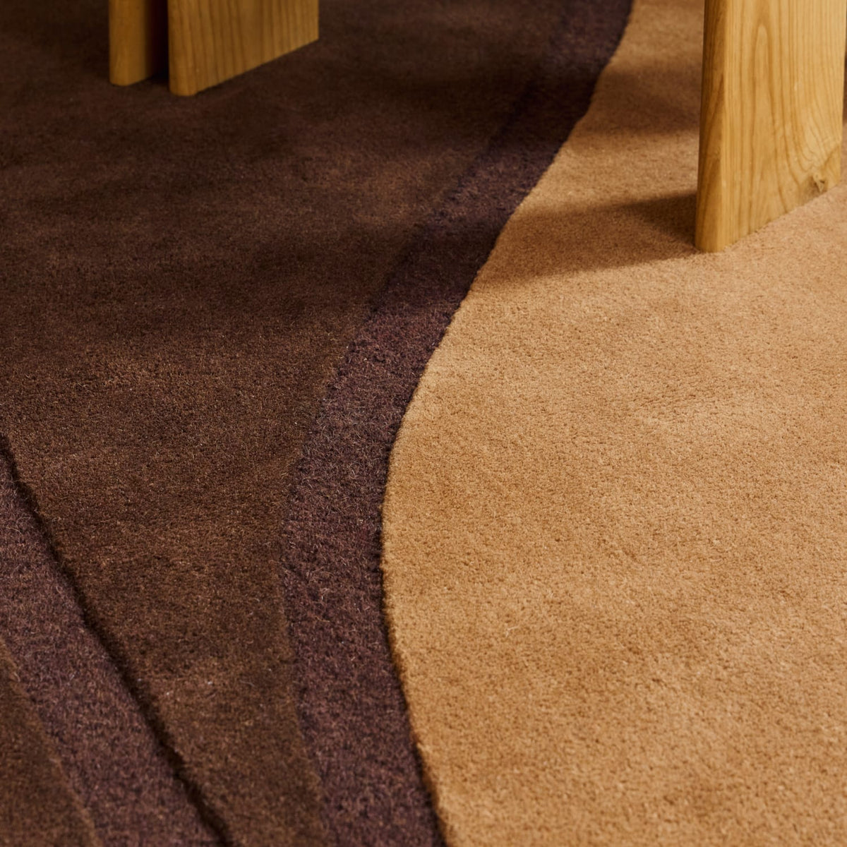 Close-up of a textured area rug with a curved organic pattern in dark brown, deep plum purple, and warm tan, with the legs of light wood furniture visible at the top.