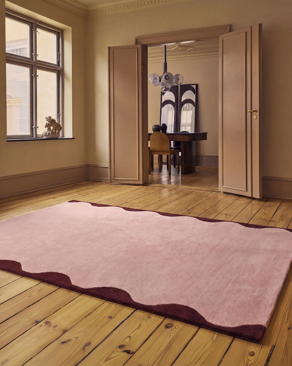 A large rectangular area rug in dusty pink with a dark burgundy wavy scalloped border, laid out on a light wood plank floor in a warm, elegant room with beige walls and open wooden doors.
