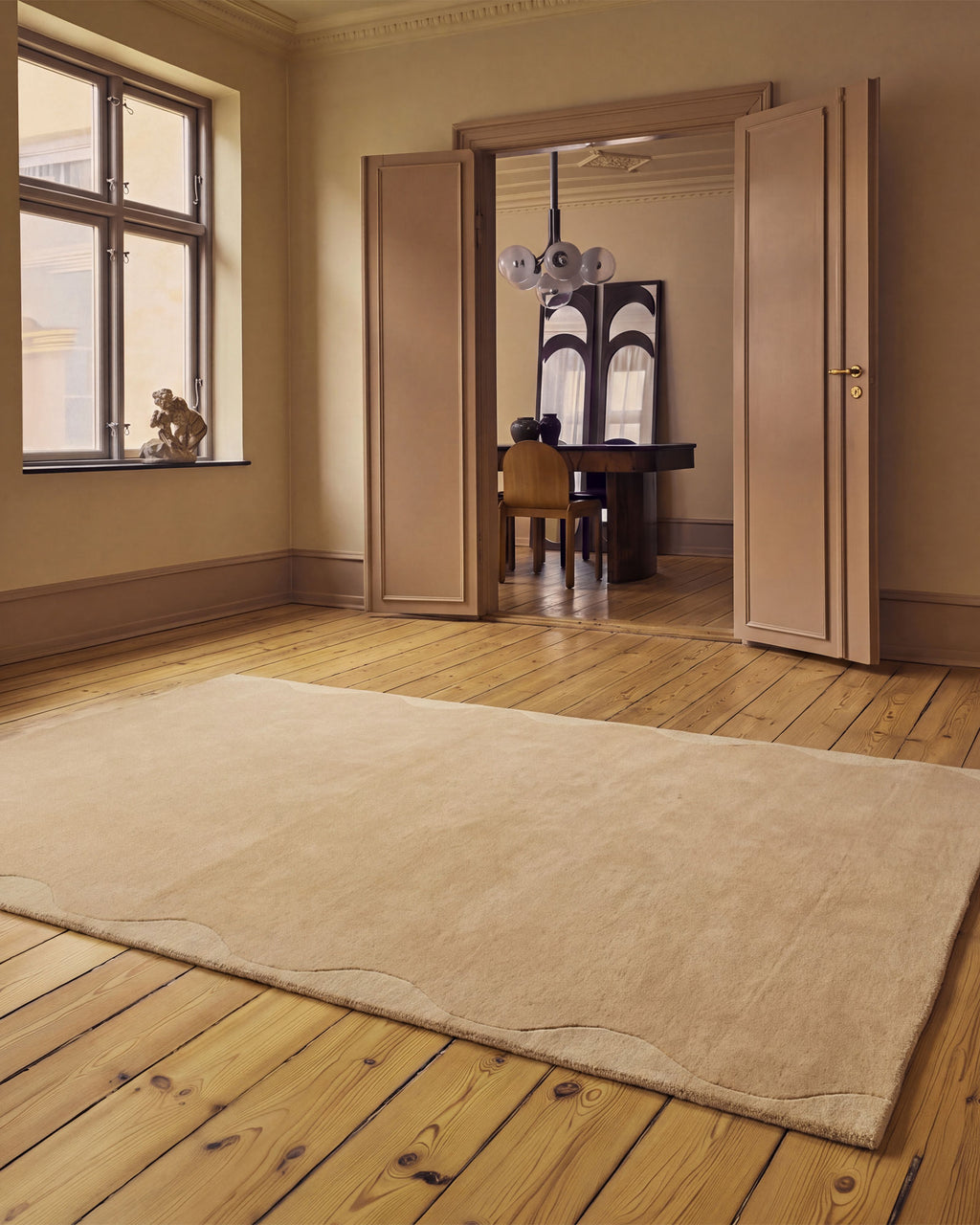 A large beige area rug laid out on a light wood plank floor in a warm, elegant room with open double doors leading to an adjacent room.