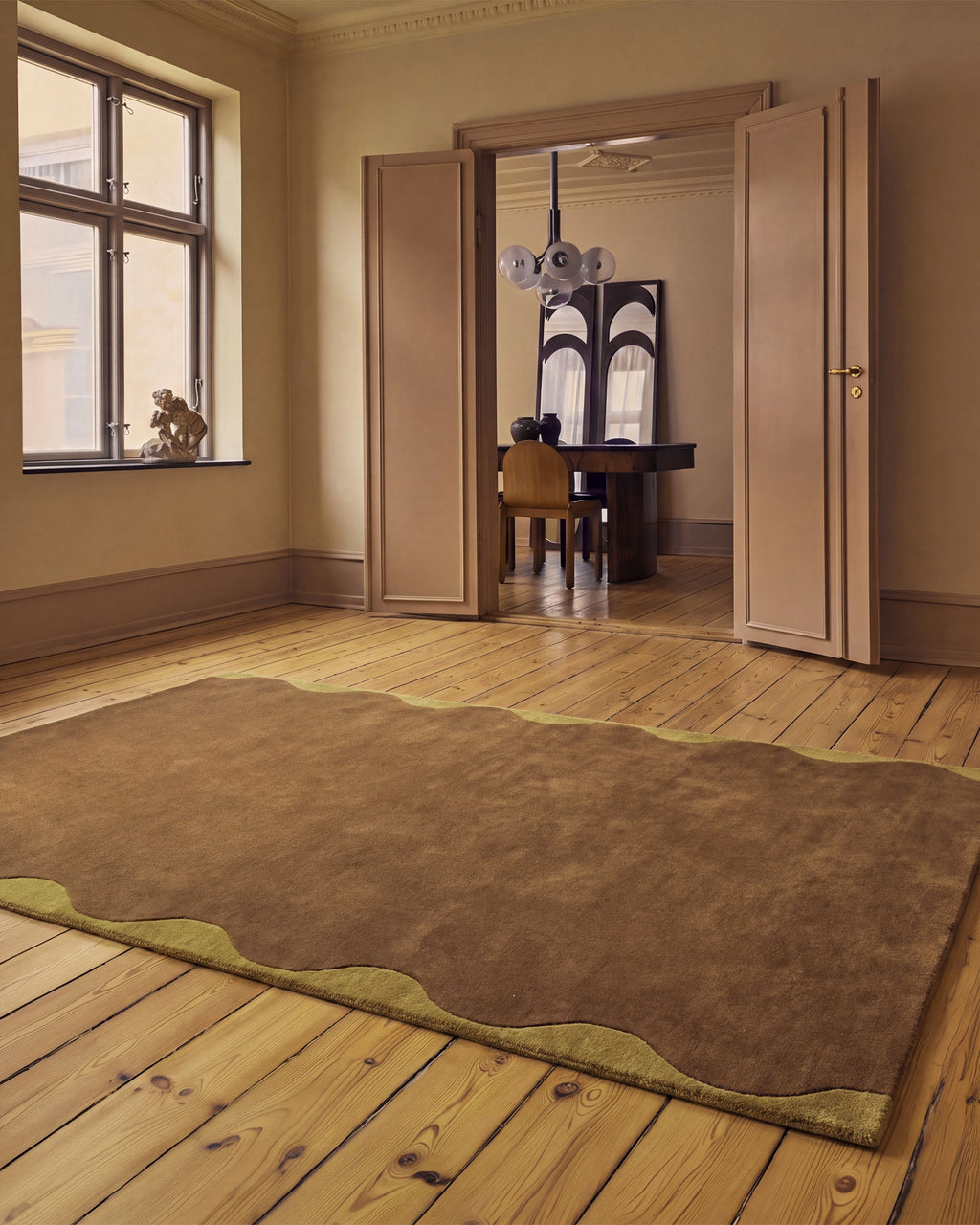 A large brown area rug with a wavy scalloped edge and olive-green trim, laid out on a light wood plank floor in a warm, elegant room. Through open double doors, a dining area with a dark table and chair is visible in the background.
