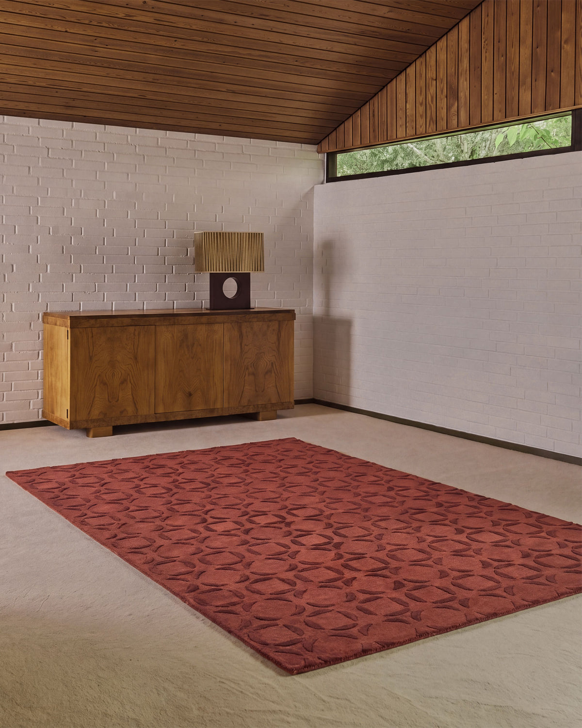 A rectangular area rug with a repeating geometric medallion pattern in rusty red and dark brown, displayed on a light beige carpet in a modern room with a wood-paneled ceiling, white brick walls, and a wooden sideboard.