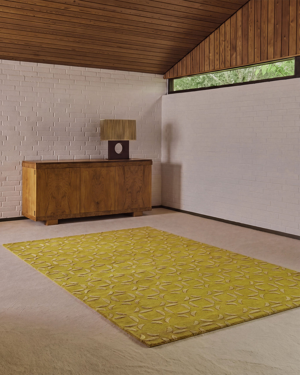 A yellow area rug with a geometric leaf pattern is displayed on a light beige carpet in a modern room with a wood-paneled ceiling, white brick wall, and a wooden sideboard.