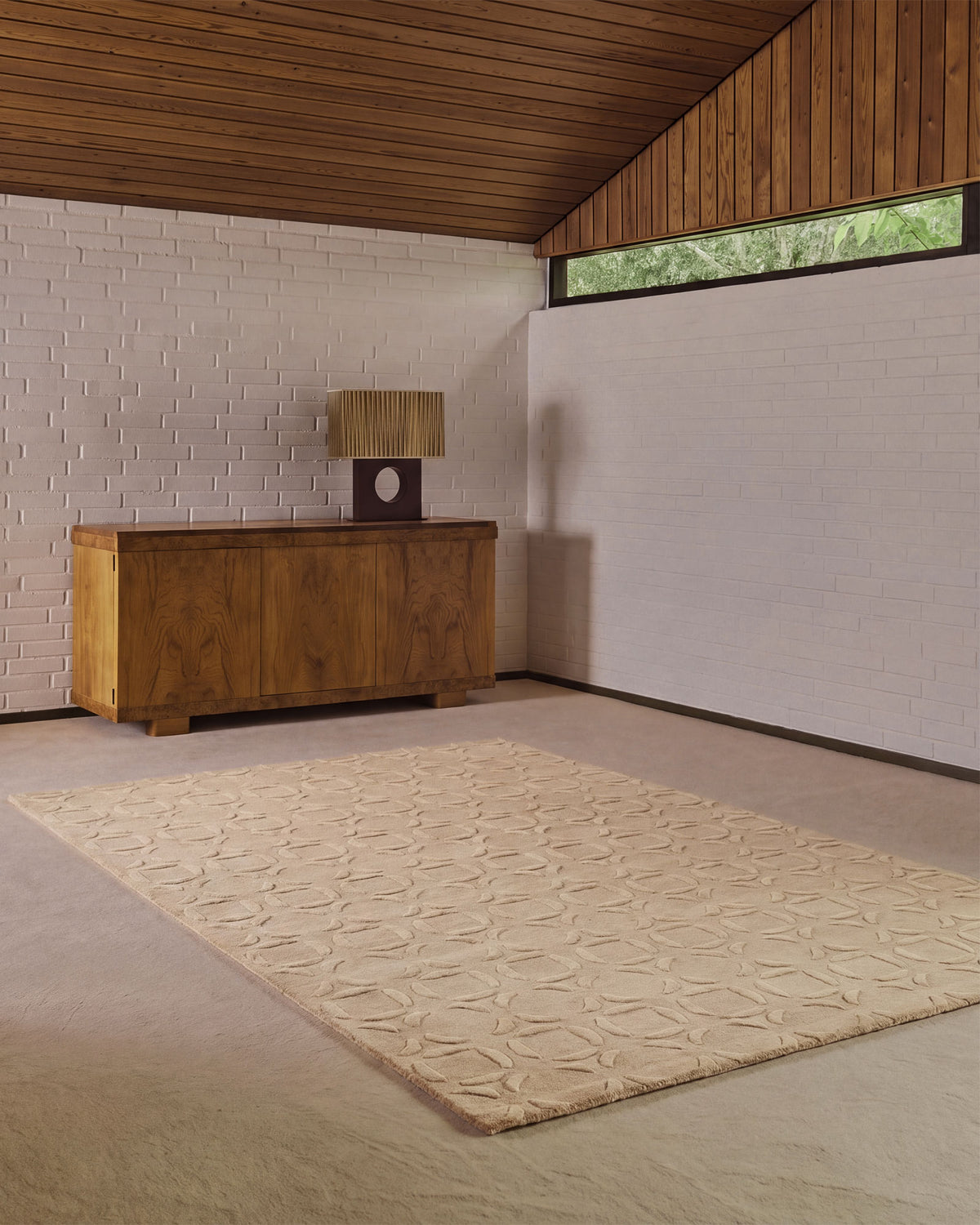 A large beige area rug with a raised geometric leaf pattern, displayed in a modern room with a wood-paneled ceiling, white brick walls, and a wooden sideboard.