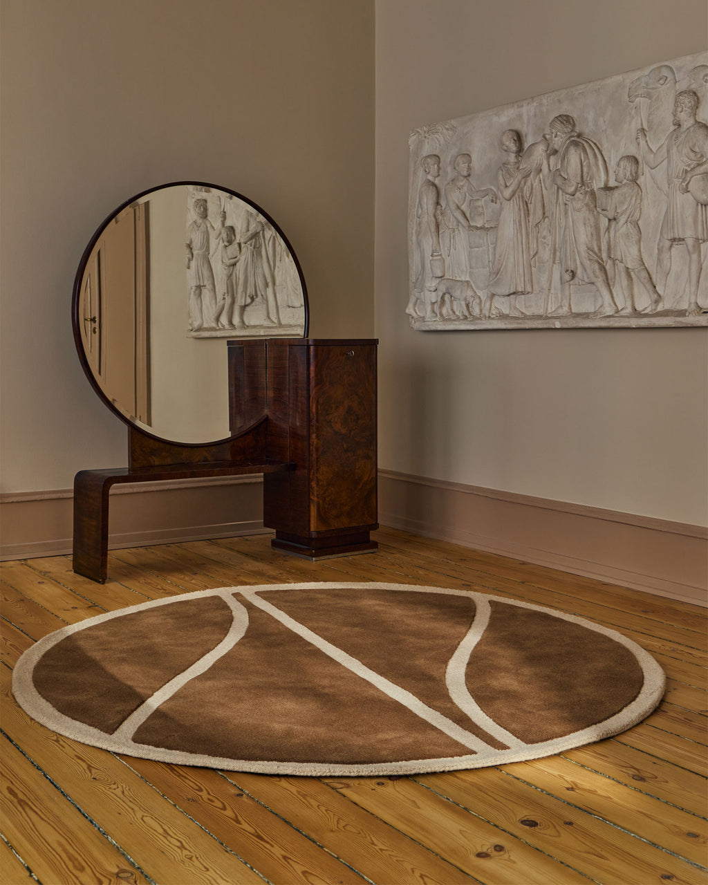 A round area rug with a brown basketball court design and cream lines, styled on a wooden floor in a room with a wooden console table, round mirror, and a white relief sculpture on the wall.