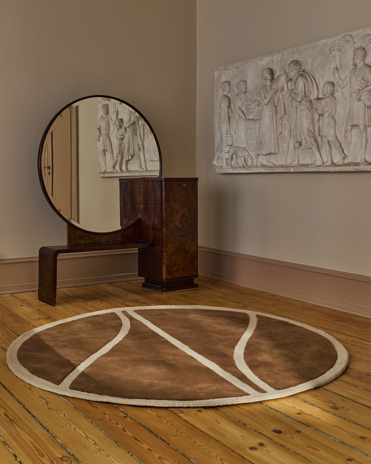 A round area rug with a brown basketball court design and cream lines, styled on a wooden floor in a room with a wooden console table, round mirror, and a white relief sculpture on the wall.