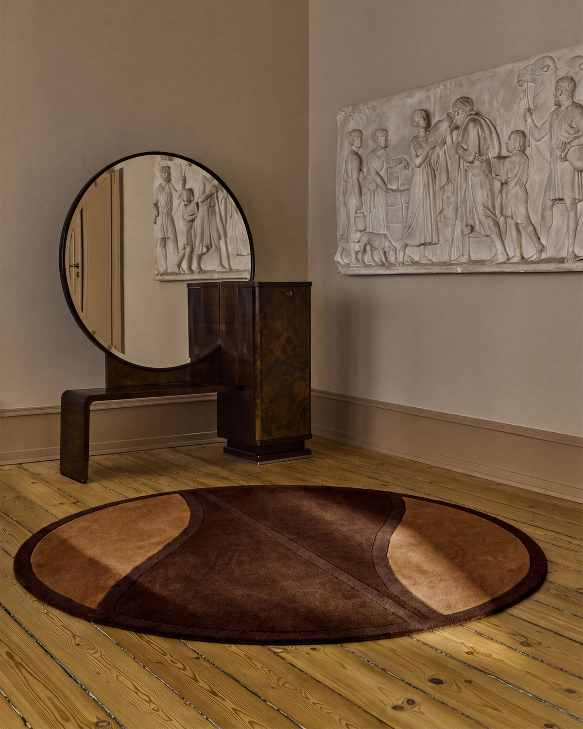 A round area rug in shades of brown and tan is styled on a light wood floor in a room setting, with a dark wooden console table and a round mirror featuring a classical relief sculpture reflection.