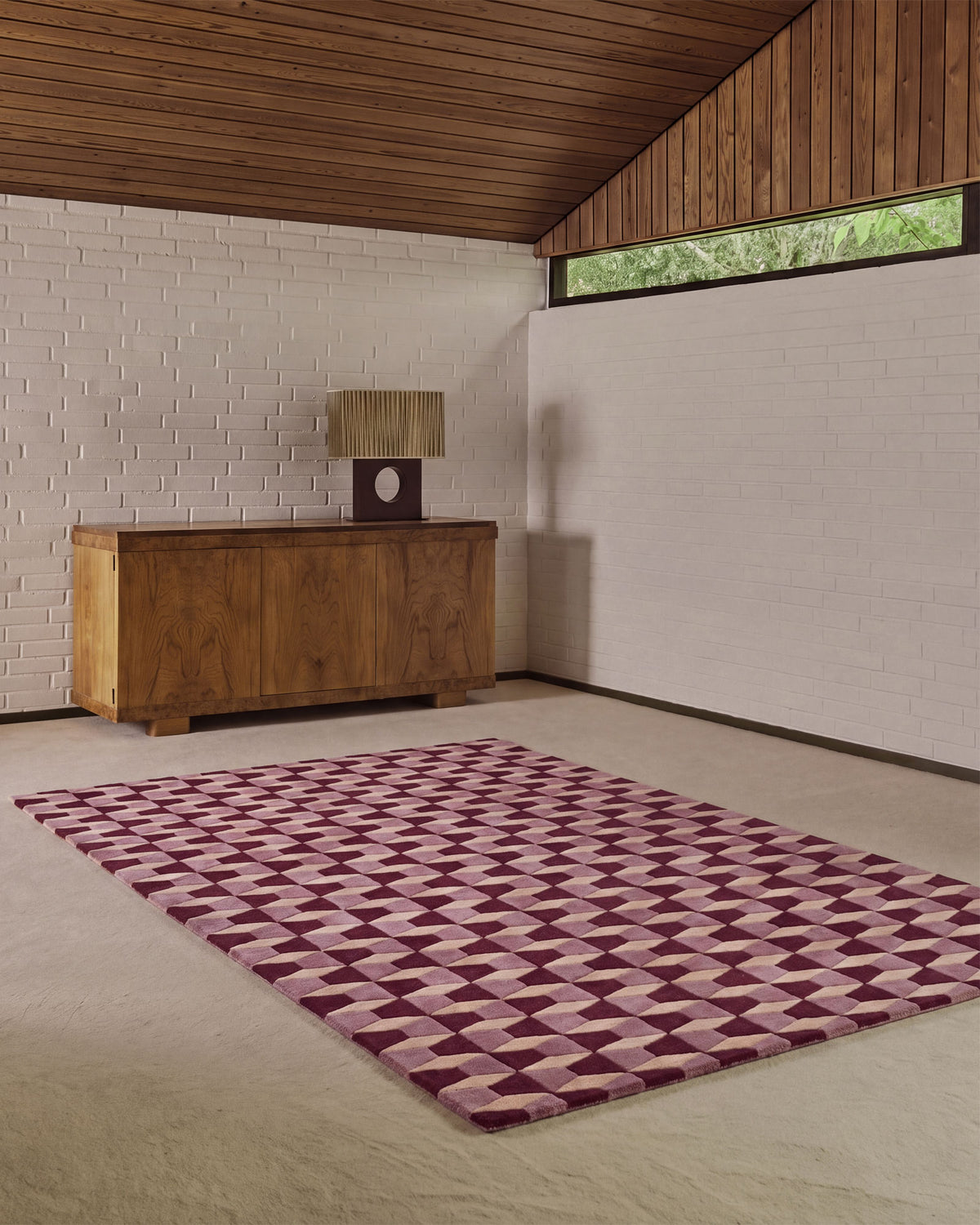 A rectangular area rug with a geometric 3D-effect pattern in shades of purple and pink, displayed on a light beige floor in a modern room with a wood-paneled ceiling and white brick wall.