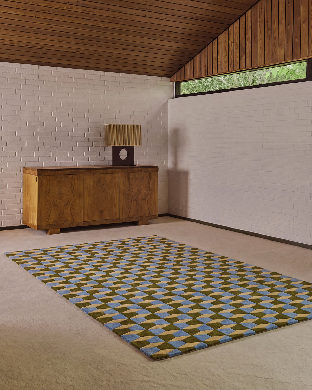 A geometric area rug with a blue, olive green, and beige diamond pattern, displayed on a light beige floor in a modern room with a wood-paneled ceiling and white brick walls.