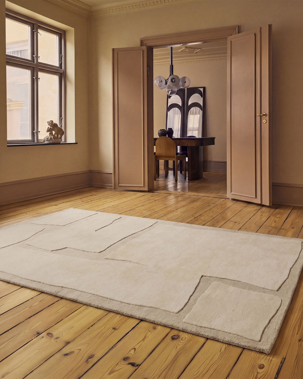 A large rectangular area rug in a warm off-white or cream color with a patchwork design, displayed on a light wood plank floor in a beautifully styled room with beige walls and open wooden doors.
