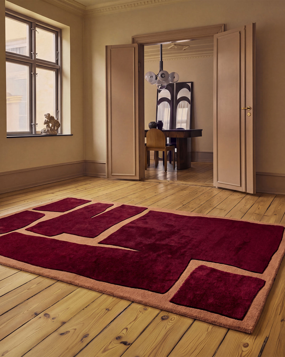 A plush rectangular area rug with an abstract geometric pattern in deep burgundy and tan, displayed on a light wood plank floor in a classic room with beige walls and open doors.