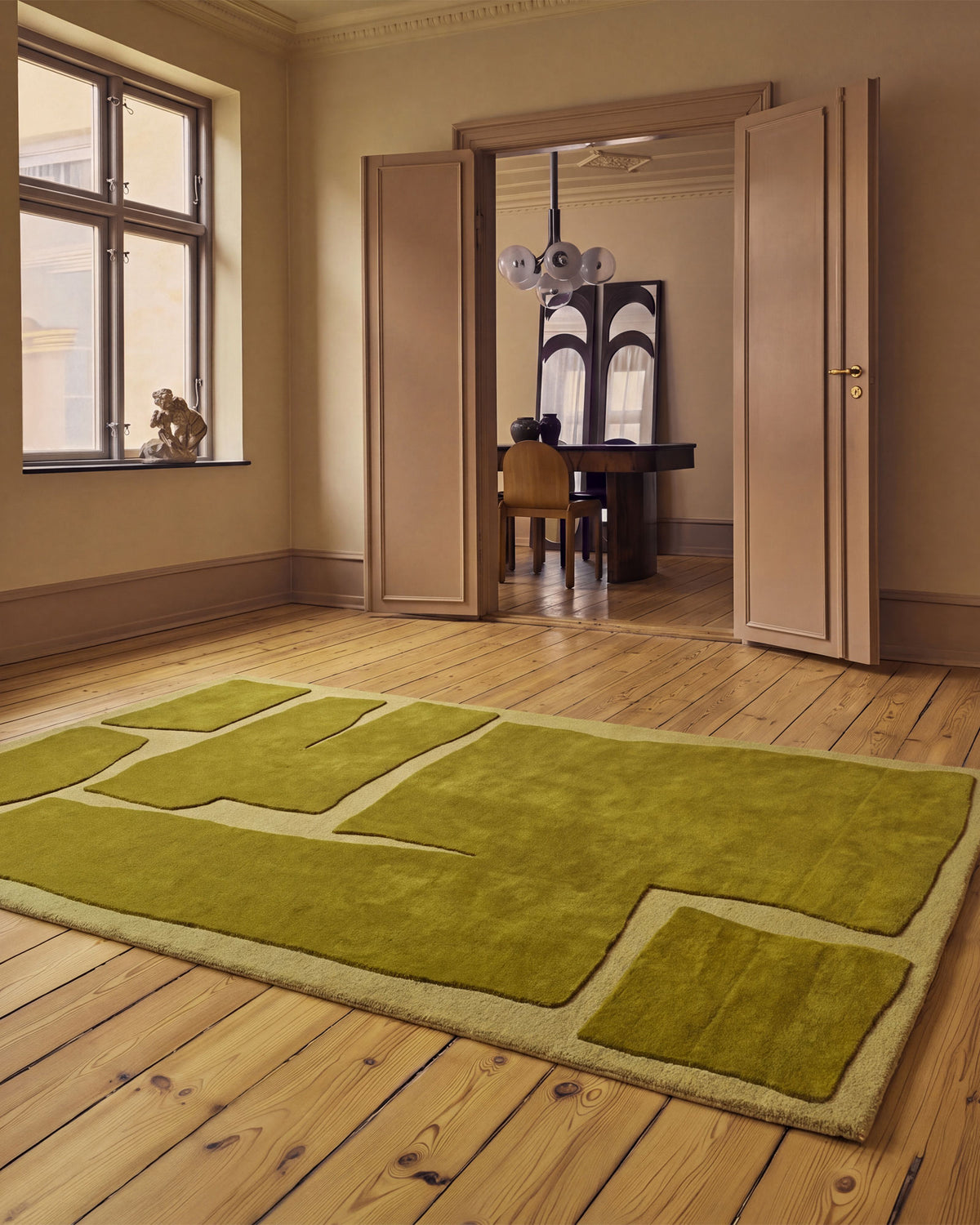 An olive green area rug and abstract geometric patchwork design, displayed on a light wood floor in a classic room with wooden paneling and open doors leading to a desk area.