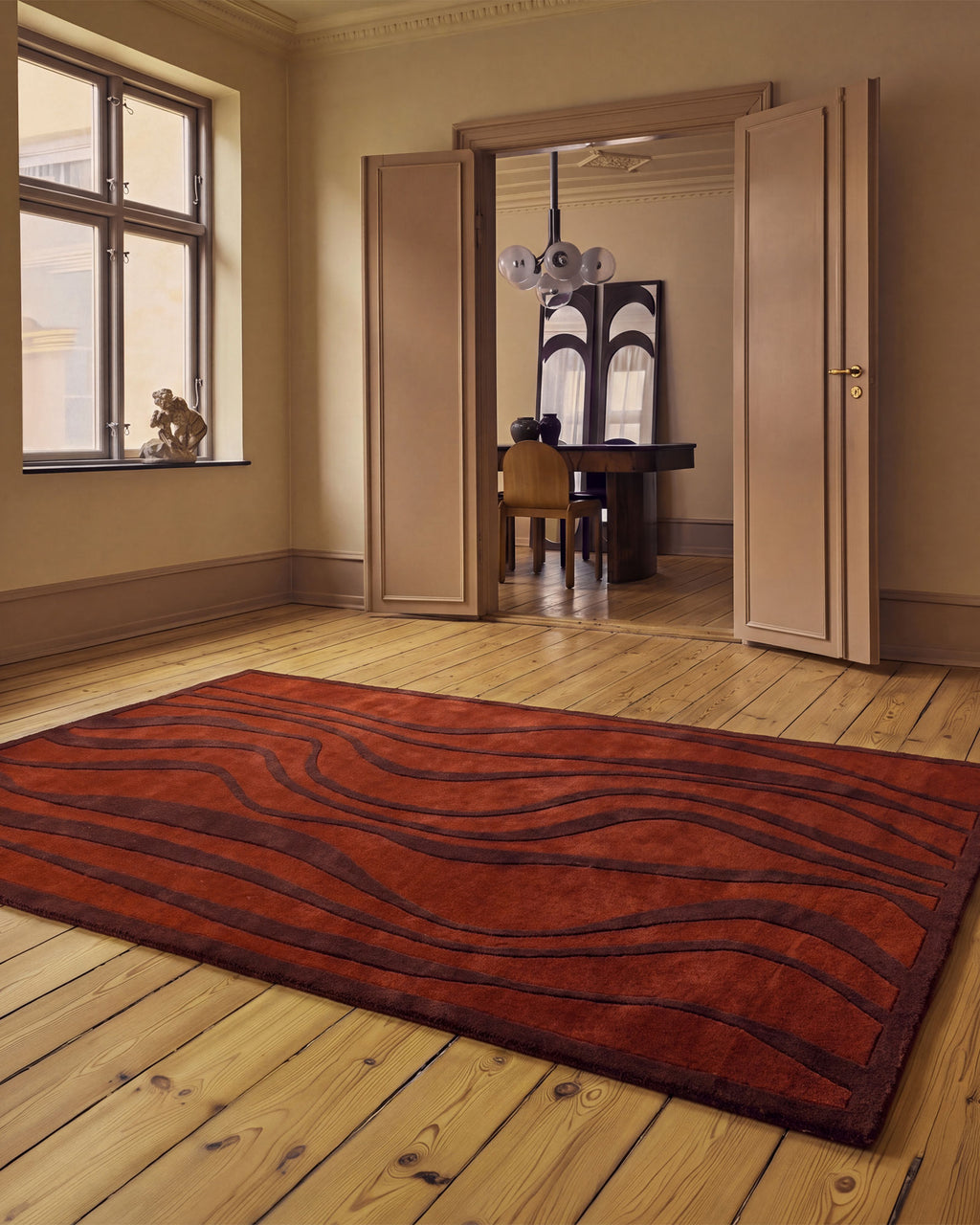 A large rectangular area rug with a wavy, organic dark brown pattern on a rust-red background, displayed on a light wood plank floor in a classic room with beige walls and an open doorway leading to another room.