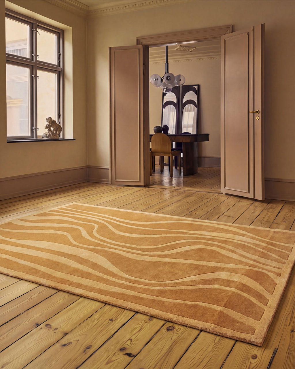 A large rectangular area rug with wavy tan and cream stripes lies on a light wood floor in a warm, elegant room with open doors leading to a dining area.