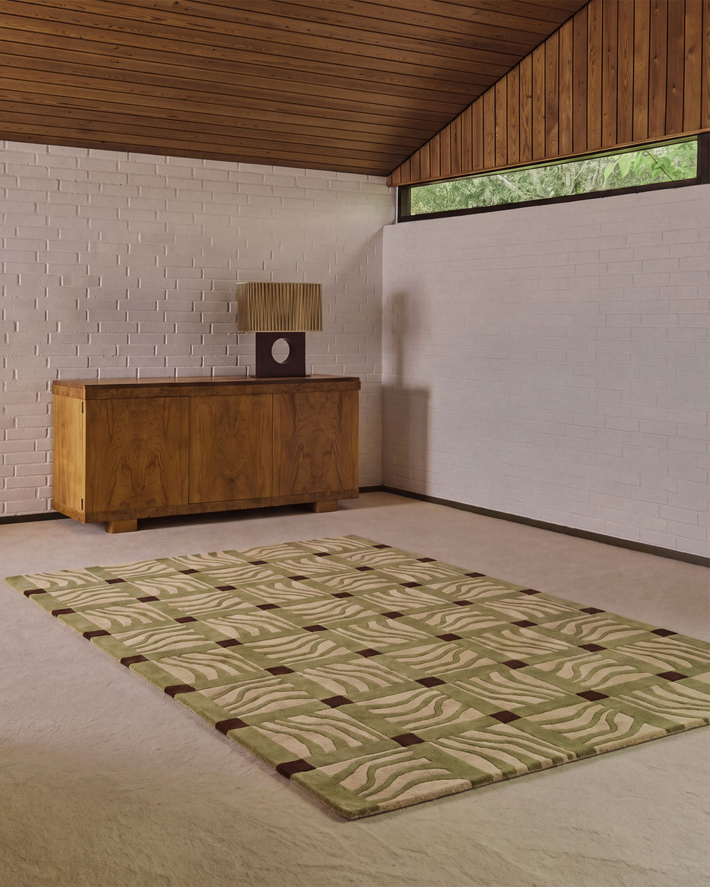A rectangular area rug with a beige and olive green wavy geometric pattern and dark brown grid accents, displayed on a light beige carpet in a modern room with a wooden credenza and white brick wall.