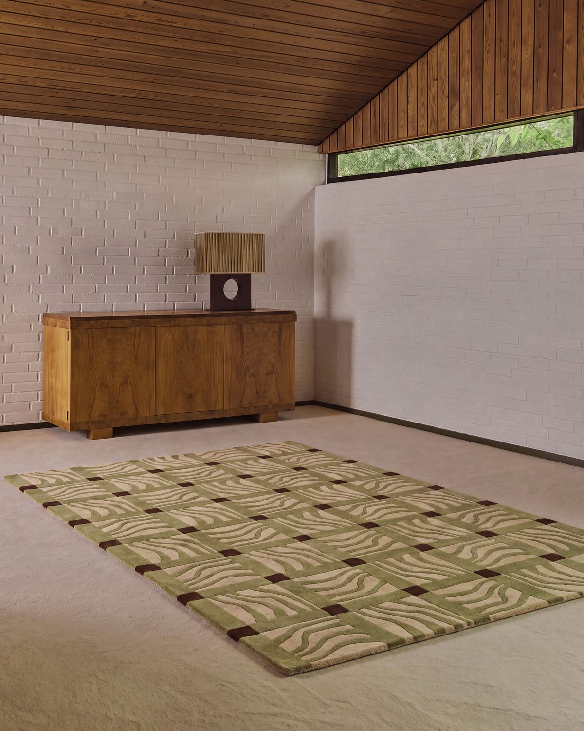 A rectangular area rug with a beige and olive green wavy geometric pattern and dark brown grid accents, displayed on a light beige carpet in a modern room with a wooden credenza and white brick wall.