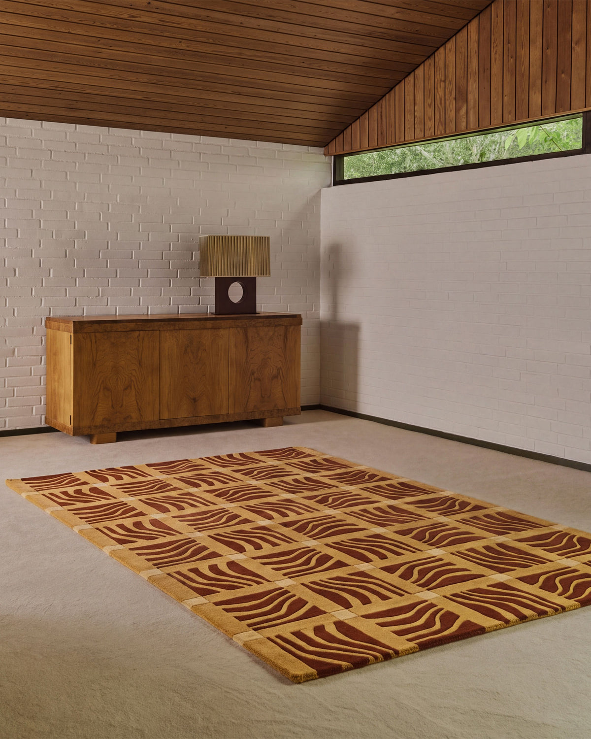 A rectangular area rug with a geometric wavy pattern in brown and beige, displayed on a light-colored floor in a modern room with a wooden credenza and white brick wall.